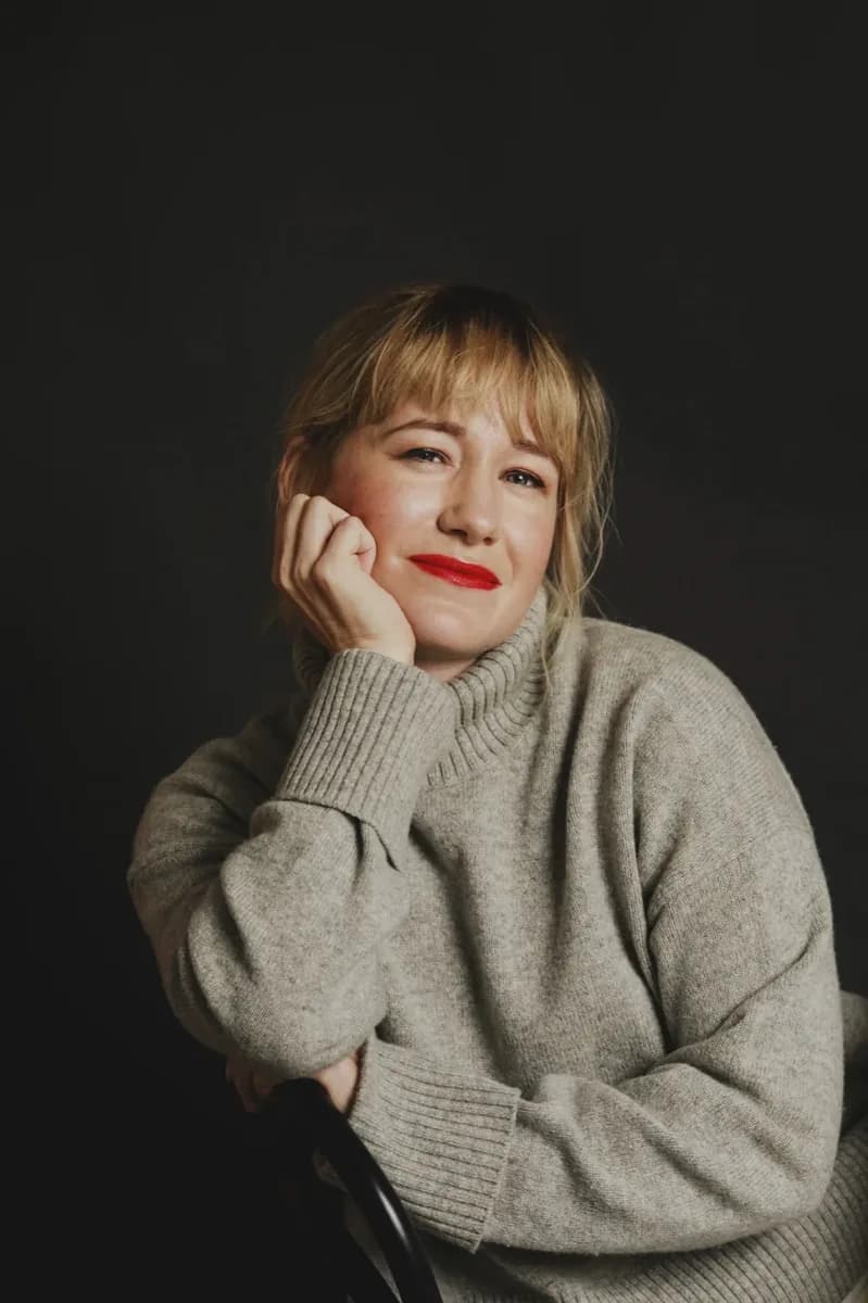 Linda Liukas in a warm grey sweater, hand resting on her cheek, smiling at the camera