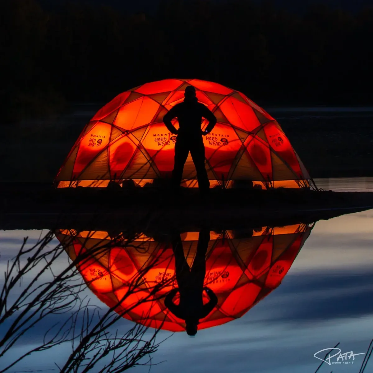 Pata Degerman silhouetted in front of a glowing dome tent reflected in still water at night